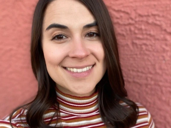 Person with shoulder length brown hair and striped turtleneck smiling with red brick background.