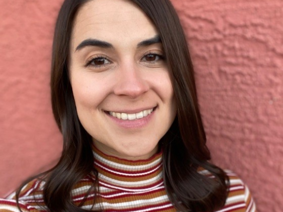 Person with shoulder length brown hair and striped turtleneck smiling with red brick background. 