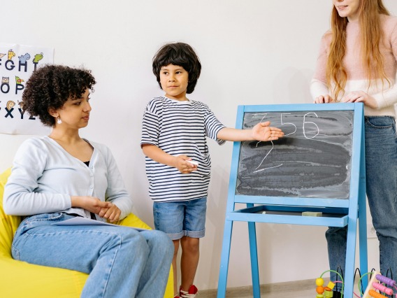 A Boy Standing Beside A Blackboard With His Teachers