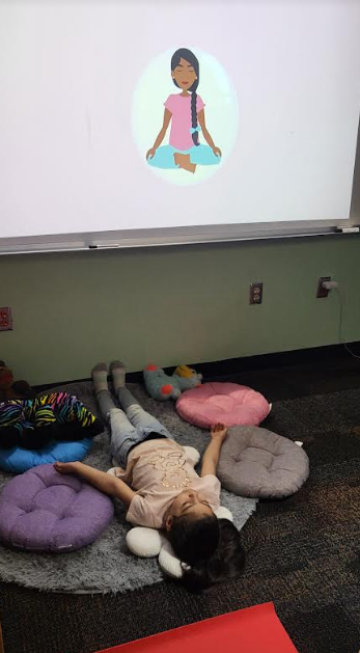 young student lying down on a grey rug surrounded by pillows watching a meditation video