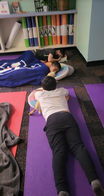 a student lying on their stomach on a purple yoga mat alongside two young students taking a nap with a blanket over them